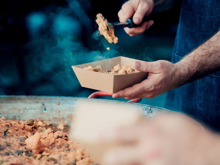 Image of A person serves a portion of rice dish from a large pan into a brown paper tray, with steam rising from the food. Another person’s hand holding a tray is visible in the foreground.