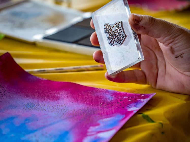 Image of A hand holds a clear stamp with a textured design above a colourful paper with pink, blue, and purple shades on a yellow table, ready for stamping.