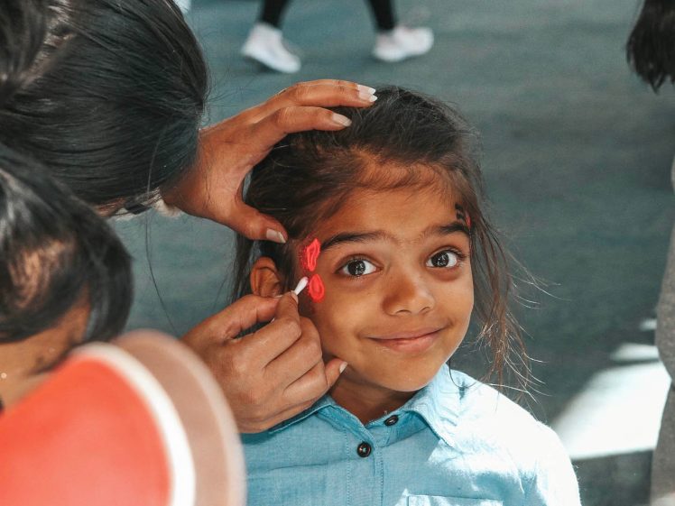 Image of A young girl smiles whilst someone paints red flowers on her cheek. She has big brown eyes and wears a blue shirt. The background is blurred with people standing and walking.