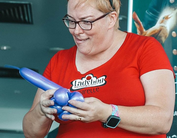 Image of A woman wearing glasses and a red shirt shapes a blue balloon into an animal. She has a pink balloon headband and a colourful apron with the words Balloon Artist Narelle.