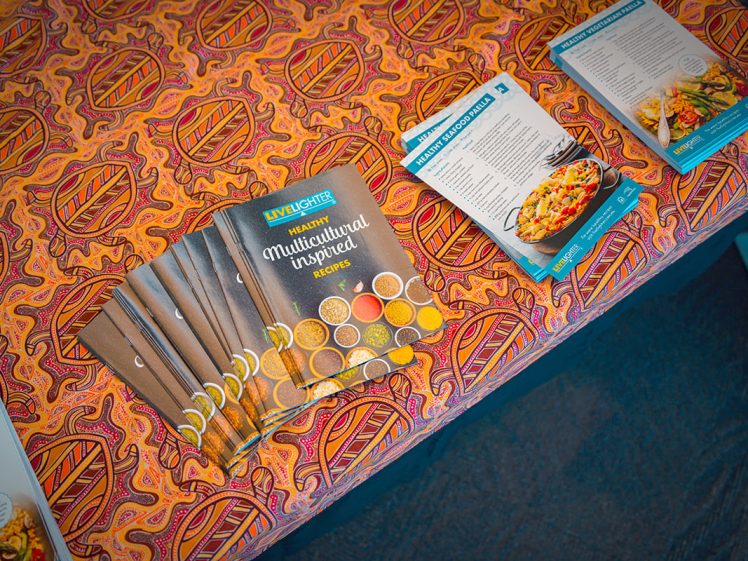 Image of A table with a colourful patterned cloth displays several copies of a booklet titled Multicultural Recipes alongside various recipe pamphlets featuring dishes and ingredients.