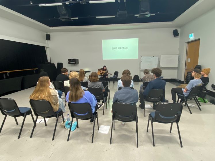 Image of A group of people sits in chairs facing a person playing guitar at the front of a room. A screen behind the performer displays the words SHOW AND SHARE. Whiteboards and a door are visible in the background.