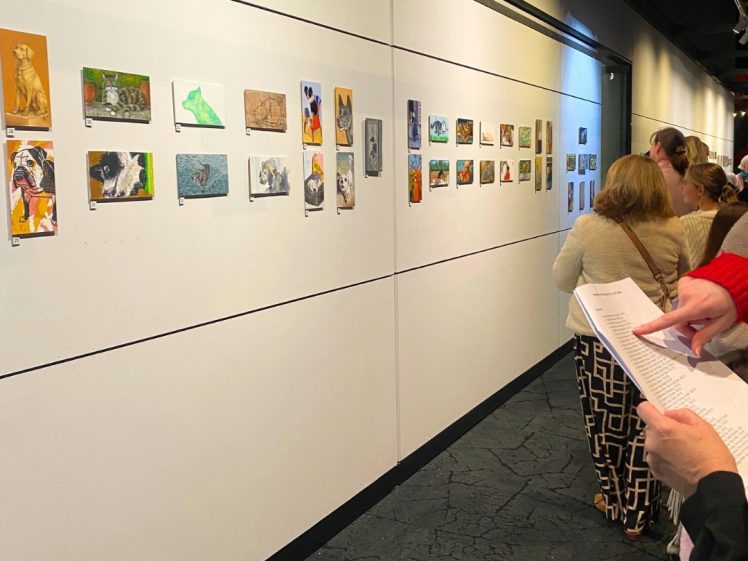 Image of People view small artworks displayed in a single row on a white gallery wall. Several individuals hold papers, possibly programmes or guides, as they observe the colourful pieces featuring various subjects.