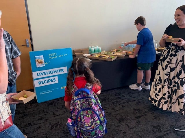 Image of A girl with a colourful rucksack stands near a table with food and drinks. A boy serves himself while a woman smiles nearby. A blue sign reads “FREE Please take one LIVELIGHTER RECIPES.”.