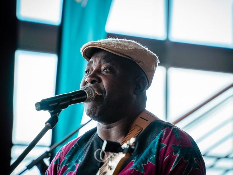 Image of A man wearing a beige flat cap and colourful shirt sings into a microphone whilst playing guitar, with a blurred background of large windows and blue curtains.