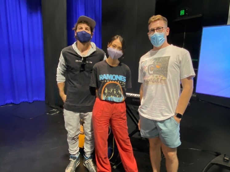 Image of Three people wearing masks stand together indoors on a dark stage with blue curtains and a keyboard behind them. One wears a hoodie, another a Ramones t-shirt and red trousers, and the third a white t-shirt and shorts.