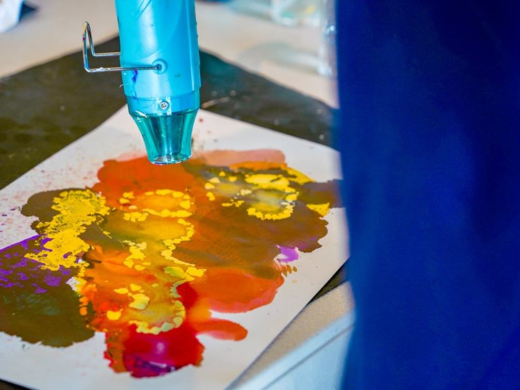 Image of A heat gun is being used to dry or manipulate colourful alcohol ink art on a piece of white paper, with vibrant hues of yellow, orange, red, and purple blending together.