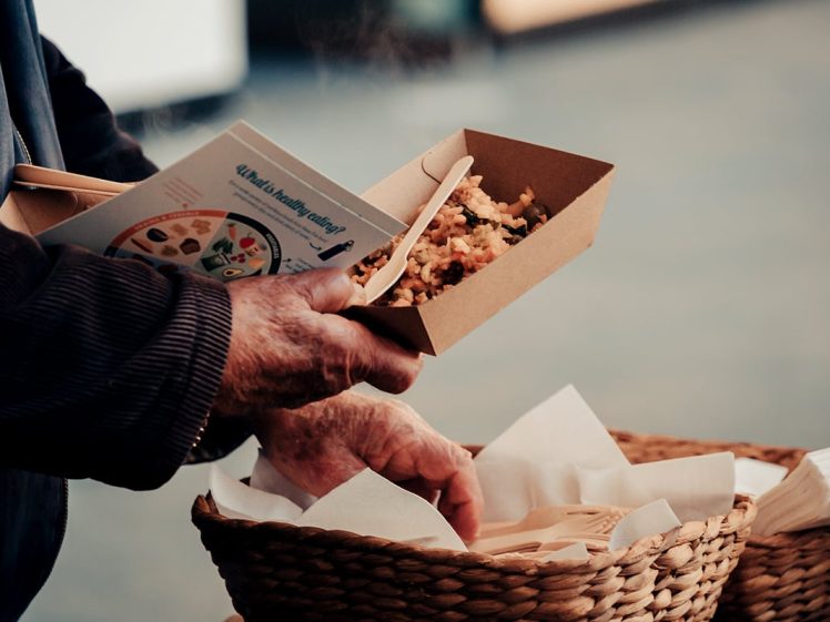 Image of A person holds a cardboard container of food and a booklet in one hand whilst reaching into a basket of wooden cutlery with the other hand. The scene suggests a casual or outdoor dining setting.