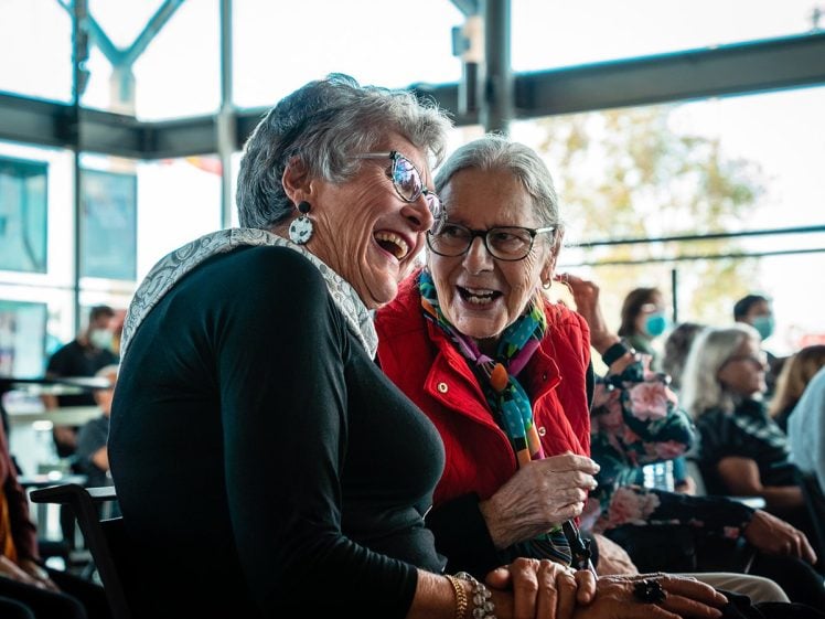 Image of Two elderly women sit close together indoors, smiling and laughing as they chat. One wears glasses, a black top, and earrings; the other wears glasses, a red jacket, and a colourful scarf. Other people are visible in the background.