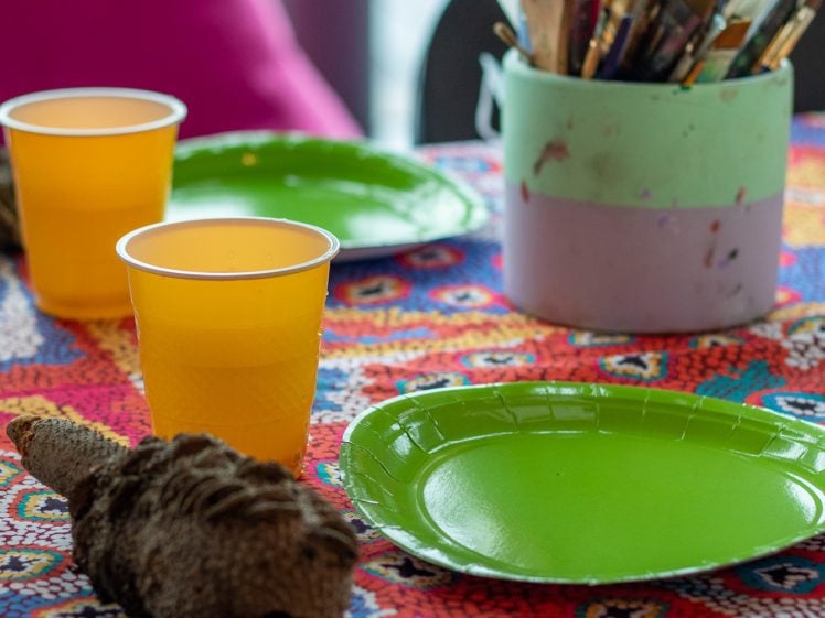 Image of A colourful table with a patterned cloth, two green plates, two orange drinks in plastic cups, a container of paintbrushes, and a woven decorative object. Bright cushions are visible in the background.