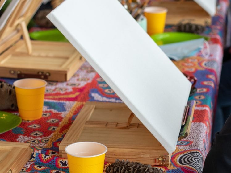 Image of A blank canvas on an easel is set on a colourful tablecloth. Nearby are a yellow plastic cup, a green plate, and a pine cone. Several people are seated at the table, preparing for a painting activity.