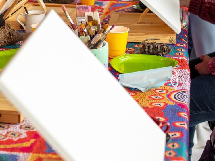 Image of A colourful table is set with blank canvases on easels, paintbrushes in cups, and yellow cups, ready for a group painting activity. People are seated round the table.