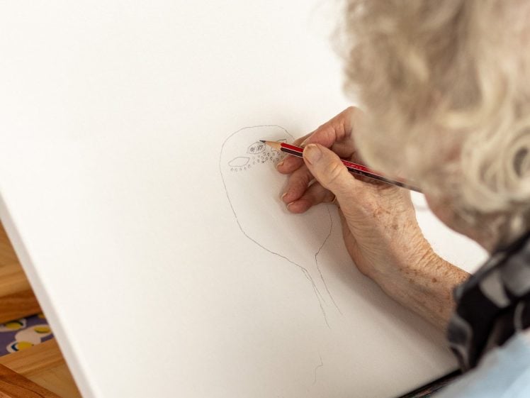 Image of An elderly person with grey hair sketches a face on a blank canvas with a pencil while seated at an easel. A colourful tablecloth is visible below the canvas.
