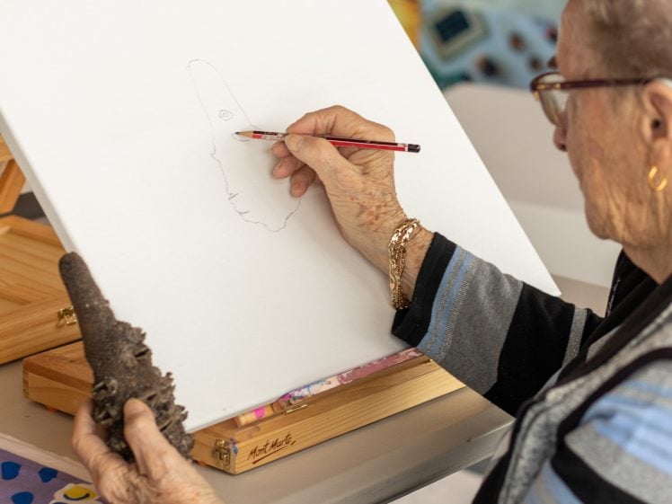 Image of An elderly woman wearing spectacles sketches a face in profile on a blank canvas, holding a pencil in one hand and a brown object in the other, while seated at a table in an art studio.