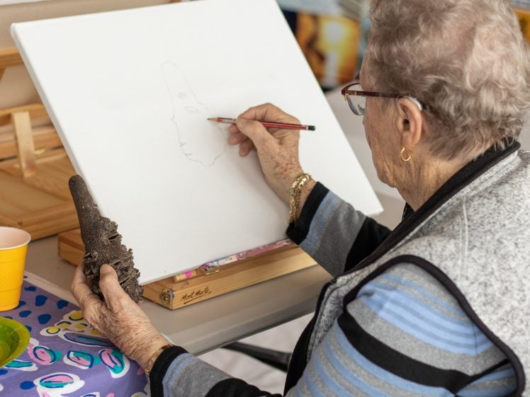 Image of An elderly woman sketches on a blank canvas with a pencil whilst holding a brown textured object in her left hand. She sits at a table with art supplies and a yellow mug nearby.