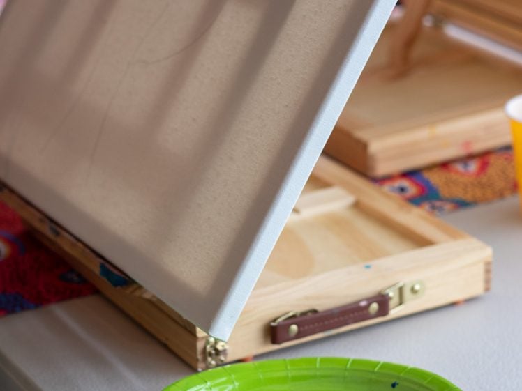 Image of A blank canvas on an easel sits next to a green paper plate with dabs of different coloured paints, including blue, green, white, brown, and red, on a table.
