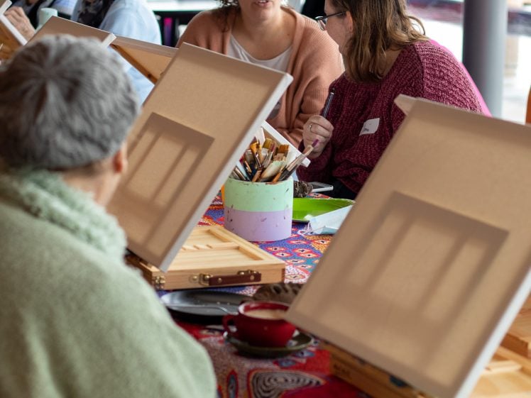 Image of Several people sit at tables with easels, painting supplies, and blank canvases. Two women in the centre talk, while others focus on their work. A large window provides natural light in the background.