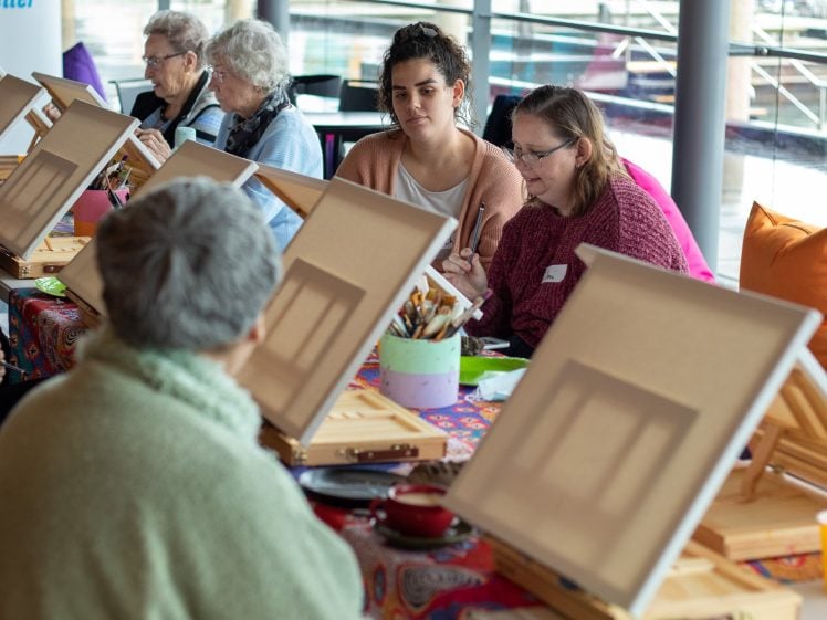 Image of A group of people of different ages sit around a table, painting on canvases set on easels. Art supplies are spread out on the table, and the atmosphere appears creative and collaborative.