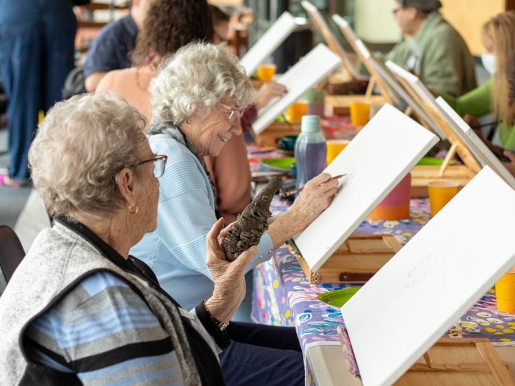 Image of A group of elderly women sit at a table with blank canvases, sketching and smiling. One woman holds a small owl sculpture as a reference. Art supplies and colourful cups are on the table.