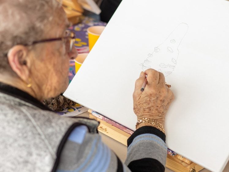 Image of An elderly person wearing spectacles sketches a cone-shaped object with circles on a blank canvas, using a pencil. The scene is bright and colourful, with art supplies visible in the background.
