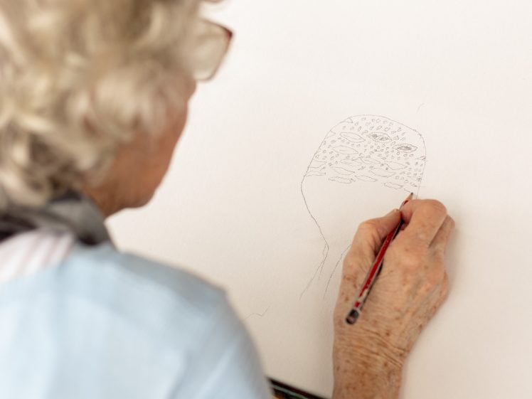 Image of An elderly person with grey hair is drawing a detailed face on white paper with a pencil, focusing on the intricate lines and shapes. The person's hand and part of their face are visible from behind.
