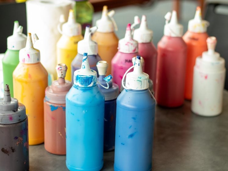Image of Several plastic squeezy bottles filled with brightly coloured paints, including blue, orange, yellow, pink, and purple, are arranged on a tabletop. Some bottles have paint splatters on their nozzles and lids.
