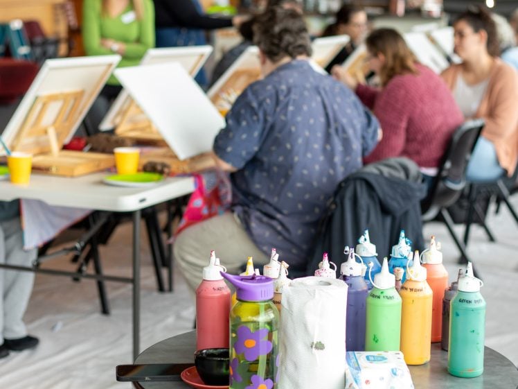 Image of A group of people sit at tables, painting on canvases in an art class. In the foreground, bottles of colourful paint and art supplies are visible on a small table. The background is out of focus.
