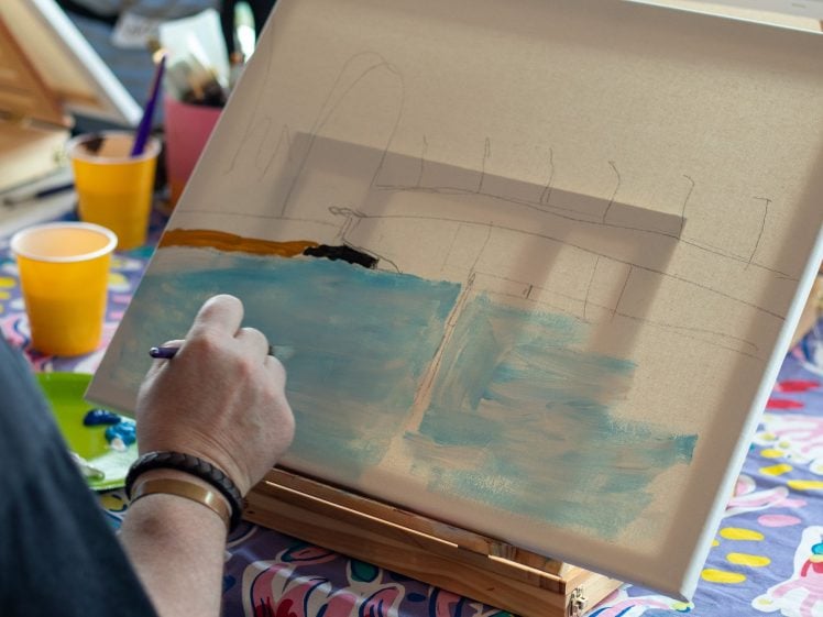 Image of A person paints a landscape with water and a bridge on a canvas. Their hand holds a brush, adding blue paint. The canvas rests on an easel, and colourful objects and cups are on the table in the background.