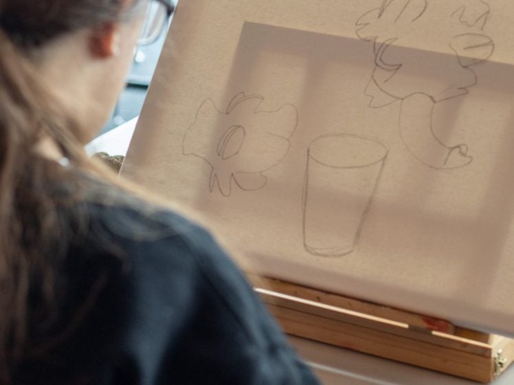 Image of A person with long hair and glasses sketches a still life featuring a flower, a leafy plant, and a glass on a canvas set on an easel. The scene is viewed from behind the artist.