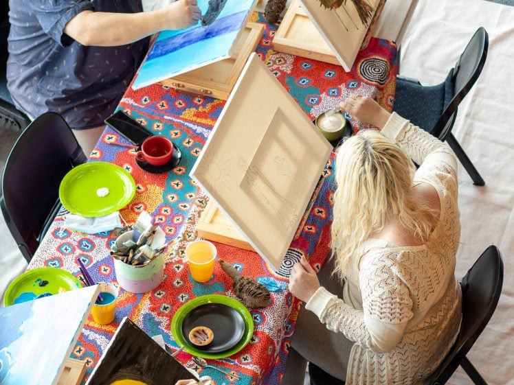 Image of Three people sit at a colourful, patterned table, each painting on canvas. The artworks feature nature themes, including trees and the Earth. Art supplies and drinks are spread across the table. The setting appears casual and creative.