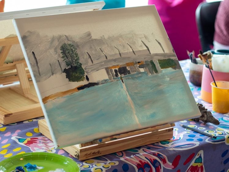Image of A canvas on an easel displays an unfinished painting of a landscape with water and trees. Art supplies, brushes, and cups are scattered on a colourful tablecloth.