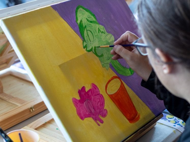 Image of A person with glasses paints a colourful still life on a canvas, featuring a green object, a pink object, and an orange cup on a split yellow and purple background. Paint supplies are visible on the table.