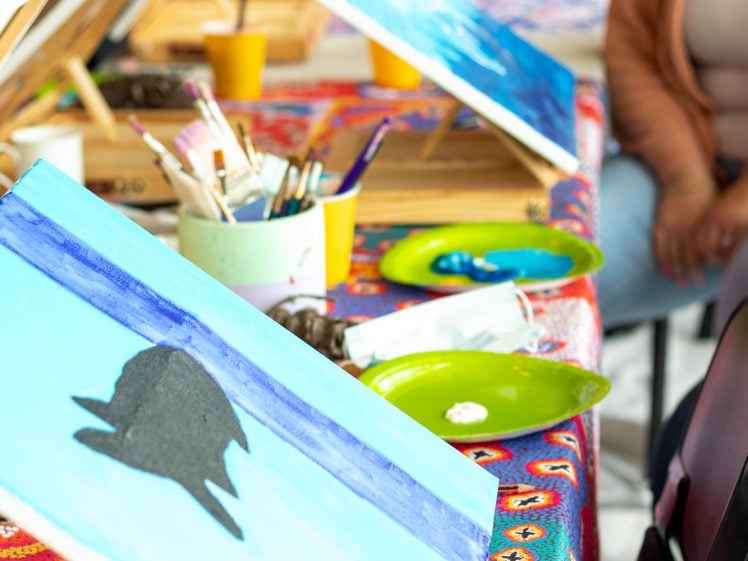 Image of A table covered with a colourful cloth holds easels with blue paintings, paintbrushes, palettes, and cups. People are seated round the table, engaged in a group painting activity.