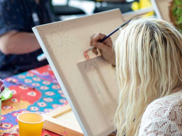 Image of A person with long blonde hair sits at a colourful table, painting a picture of a sun and a car on a canvas. Paintbrushes and art supplies are visible nearby.