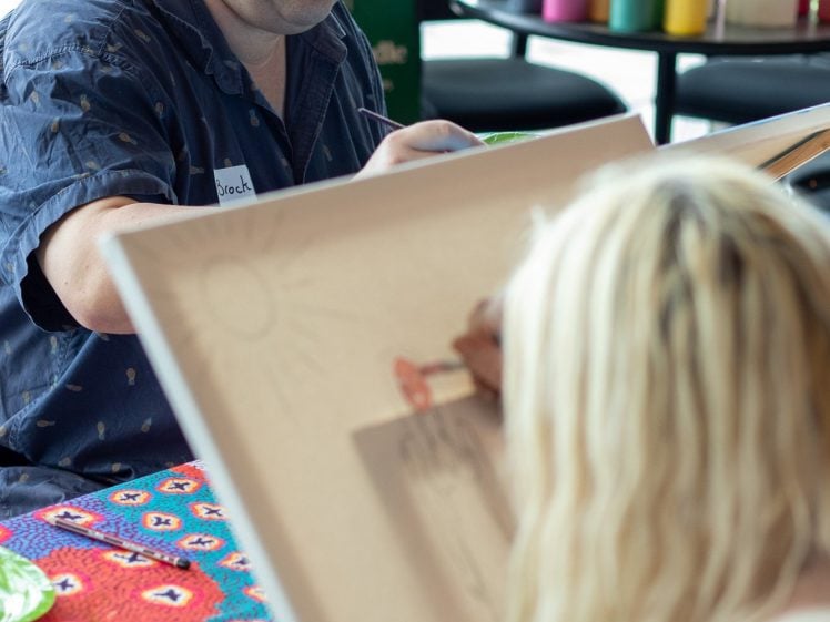 Image of A man paints at a table covered with a colourful cloth, while a woman with long blonde hair draws on a canvas in the foreground. Art supplies and paint bottles are visible in the background.
