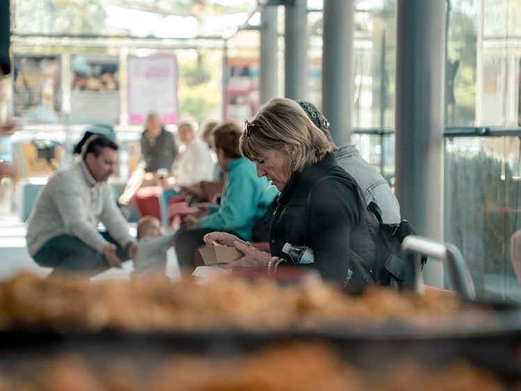 Image of A woman sits indoors at a table, focused on opening a food container. Other people are seated and talking in the background, and a blurry tray of food is visible in the foreground. Bright natural light fills the space.