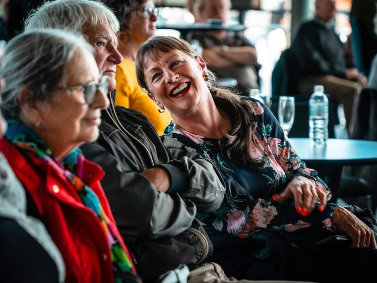 Image of A group of people sit closely together at an indoor event, smiling and laughing. The focus is on a woman in a floral top, laughing with others. Tables, drinks, and more attendees are visible in the background.