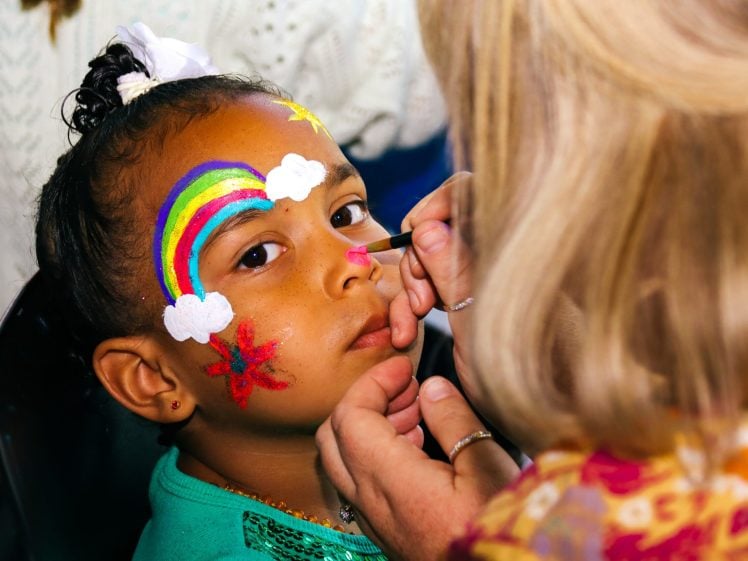 Image of A young girl with colourful face paint, including a rainbow, clouds, and stars, looks at the camera while an adult applies pink paint to her nose with a small brush.