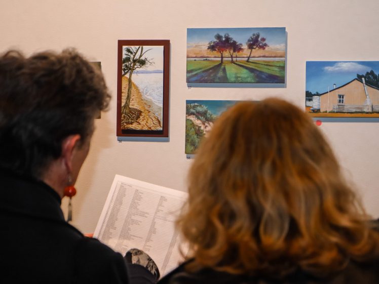 Image of Two people with their backs to the camera view a wall displaying several landscape paintings. One person holds a sheet of paper, possibly a programme or list of artworks.