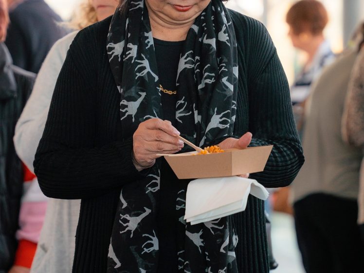 Image of A woman with short dark hair, wearing a black jumper and a scarf with white animal prints, holds a food container and fork while standing in a busy indoor setting with people in the background.