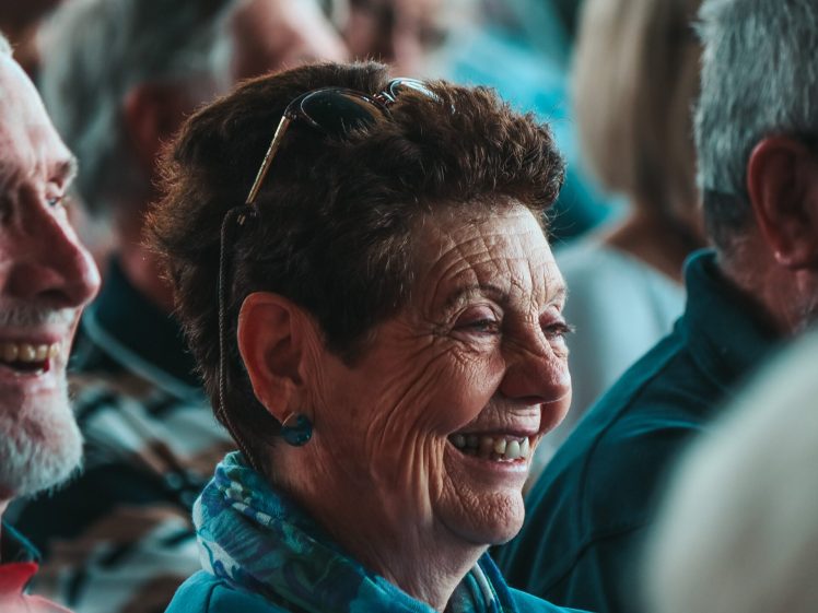 Image of A group of older adults sitting closely together, smiling and laughing. The focus is on a woman with short curly hair, glasses on her head, and a blue scarf, enjoying the moment with others around her.