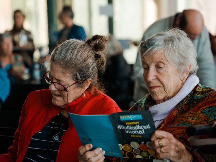 Image of Two elderly women sit together, closely reading a colourful booklet titled Healthy Multicultural Inspired Recipes in a busy indoor setting with other people in the background.