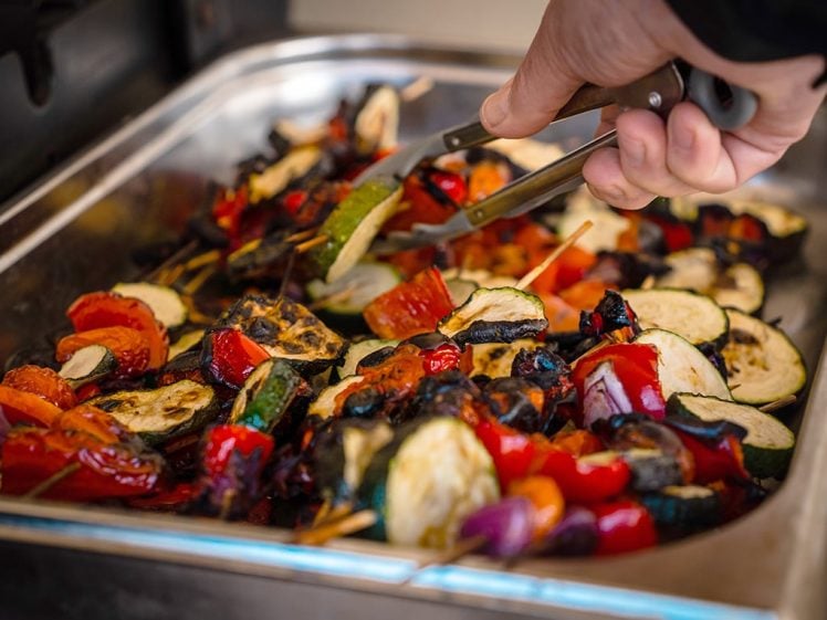 Image of A hand uses tongs to pick up grilled mixed vegetables, including courgette, peppers, and onions, from a stainless steel serving tray.