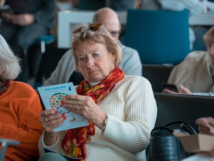 Image of An older woman wearing a white jumper and red scarf reads a colourful leaflet while sitting among other people in a waiting area. She looks focused; others are seated nearby, some using their mobiles.