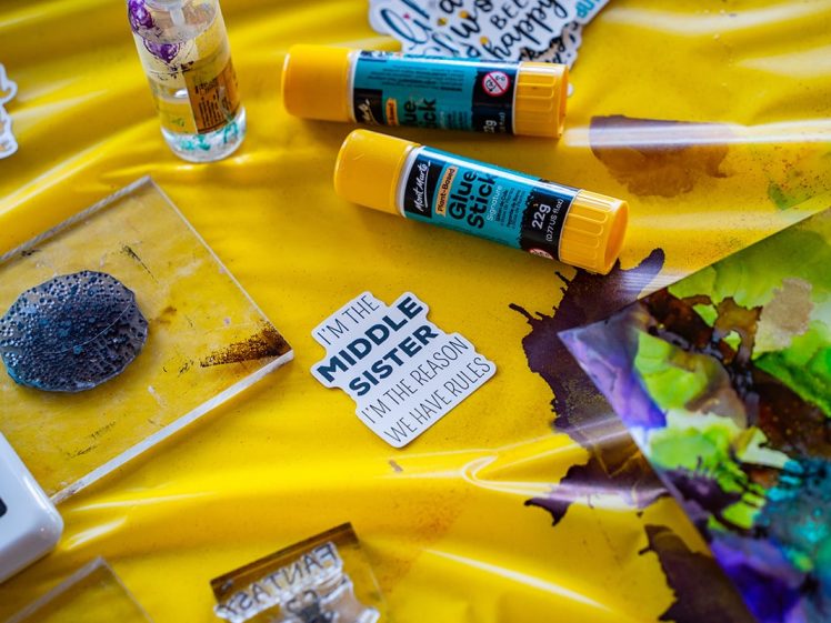 Image of A yellow craft table with two glue sticks, colourful art supplies, papers, and a sticker that reads, I'M THE MIDDLE SISTER, I'M THE REASON WE HAVE RULES.