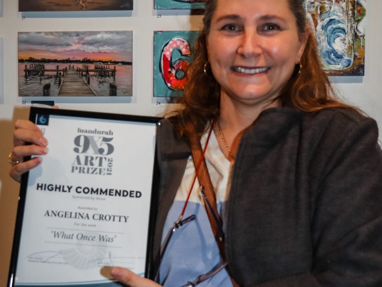 Image of A smiling woman holds a framed “Highly Commended” art award in front of a wall displaying colourful artwork, including paintings of a dog, fish, and various abstract designs.