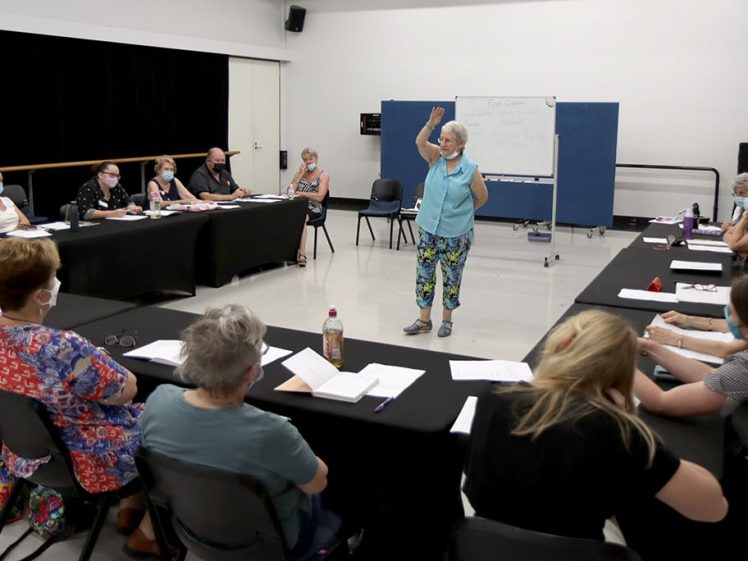 Image of A group of adults sit around tables in a classroom, listening to a woman standing and speaking in the centre. Papers and notebooks are on the tables, and a whiteboard is in the background.
