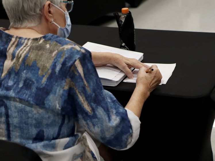 Image of An older person wearing a mask and blue-patterned shirt sits at a black table, writing on papers with a pencil. An orange-capped drink bottle is nearby. The person appears focused on their work.
