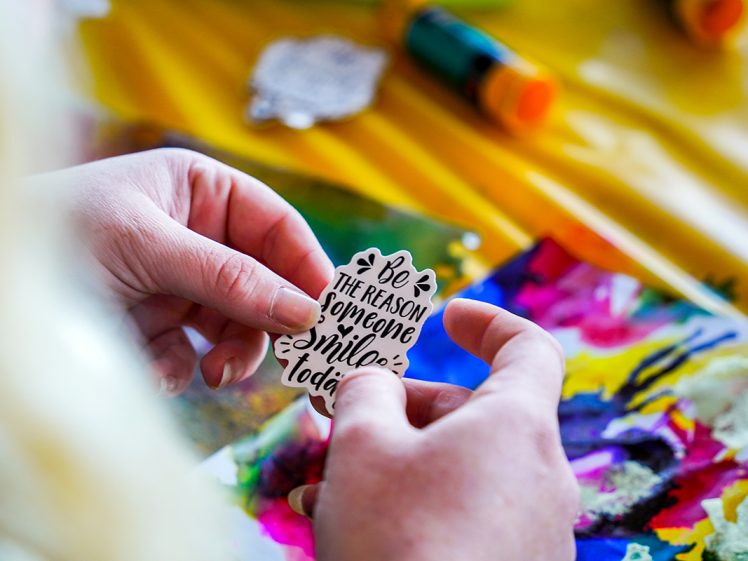 Image of A person holds a sticker that reads, Be the reason someone smiles today, with colourful art supplies and painted paper in the background.