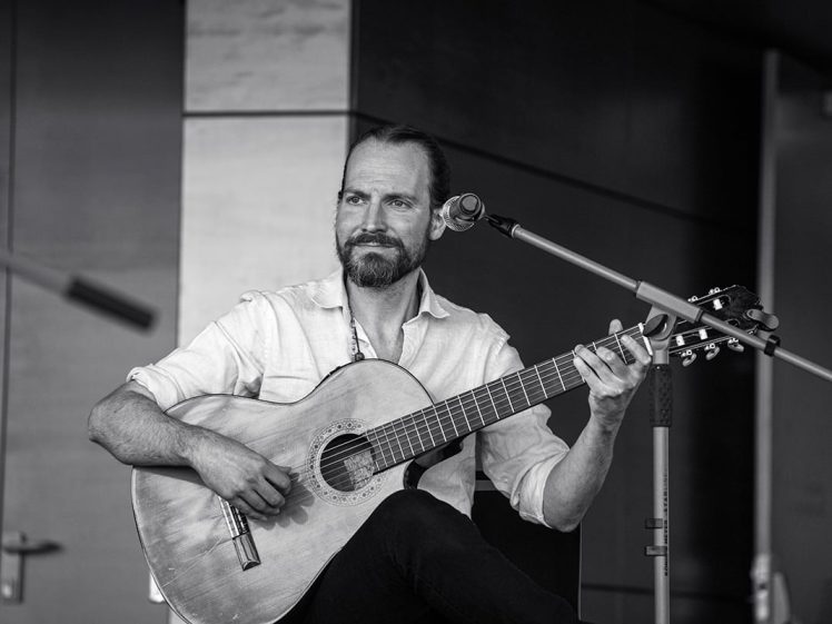 Image of A man with a beard and moustache sits on a chair, playing an acoustic guitar and looking to the side. He wears a light-coloured shirt and is positioned in front of a microphone in a modern indoor setting.